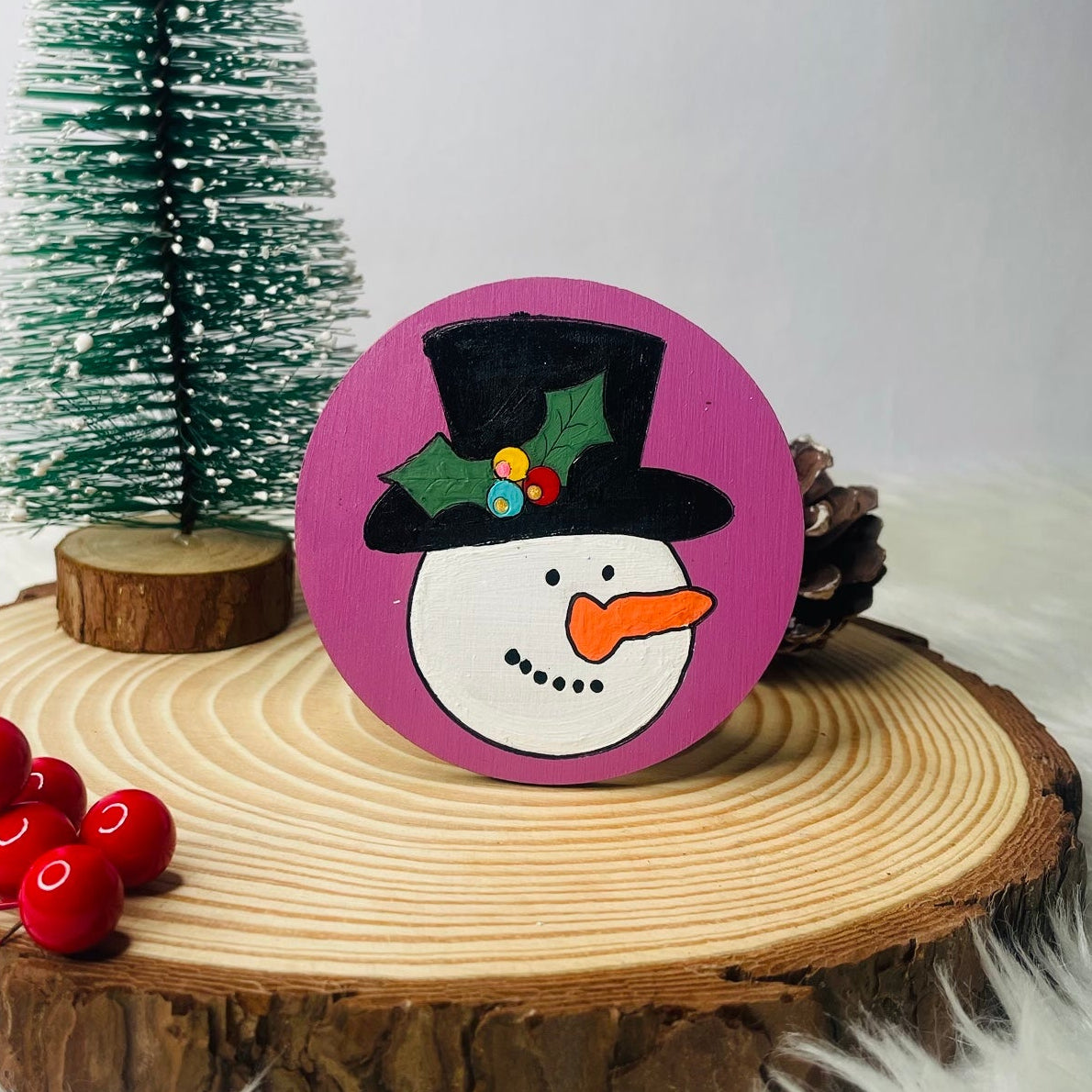 Decorative snowman ornament on a wooden slice with a small Christmas tree in the background.
