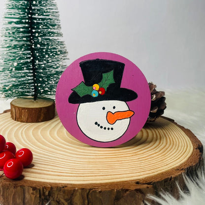 Decorative snowman ornament on a wooden slice with a small Christmas tree in the background.